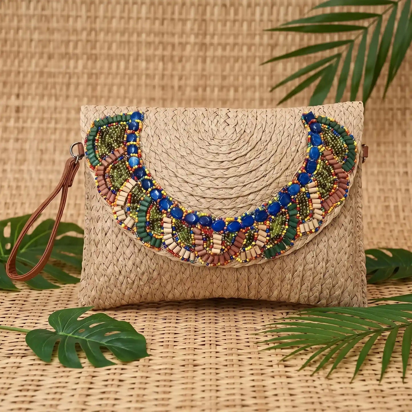 Pochette estiva in paglia intrecciata marrone chiaro con ricamo etnico di perline, posata su un tappeto di paglia con foglie tropicali - SUNTIMEMODA