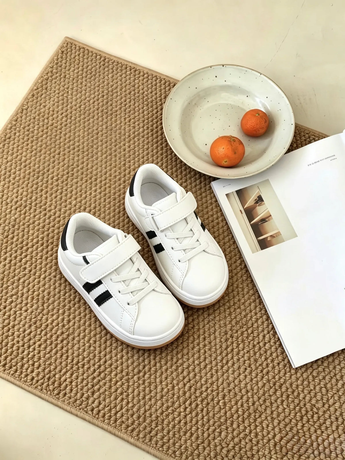 Un paio di sneakers bianche per bambini con dettagli neri e suola in gomma, ambientazione lifestyle su tappeto intrecciato. - SUNTIMEMODA