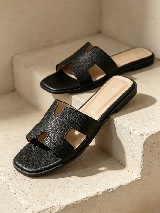 Ciabatte slide da donna nere con fascia ad H in ecopelle effetto martellato, posate su gradini in pietra chiara - SUNTIMEMODA