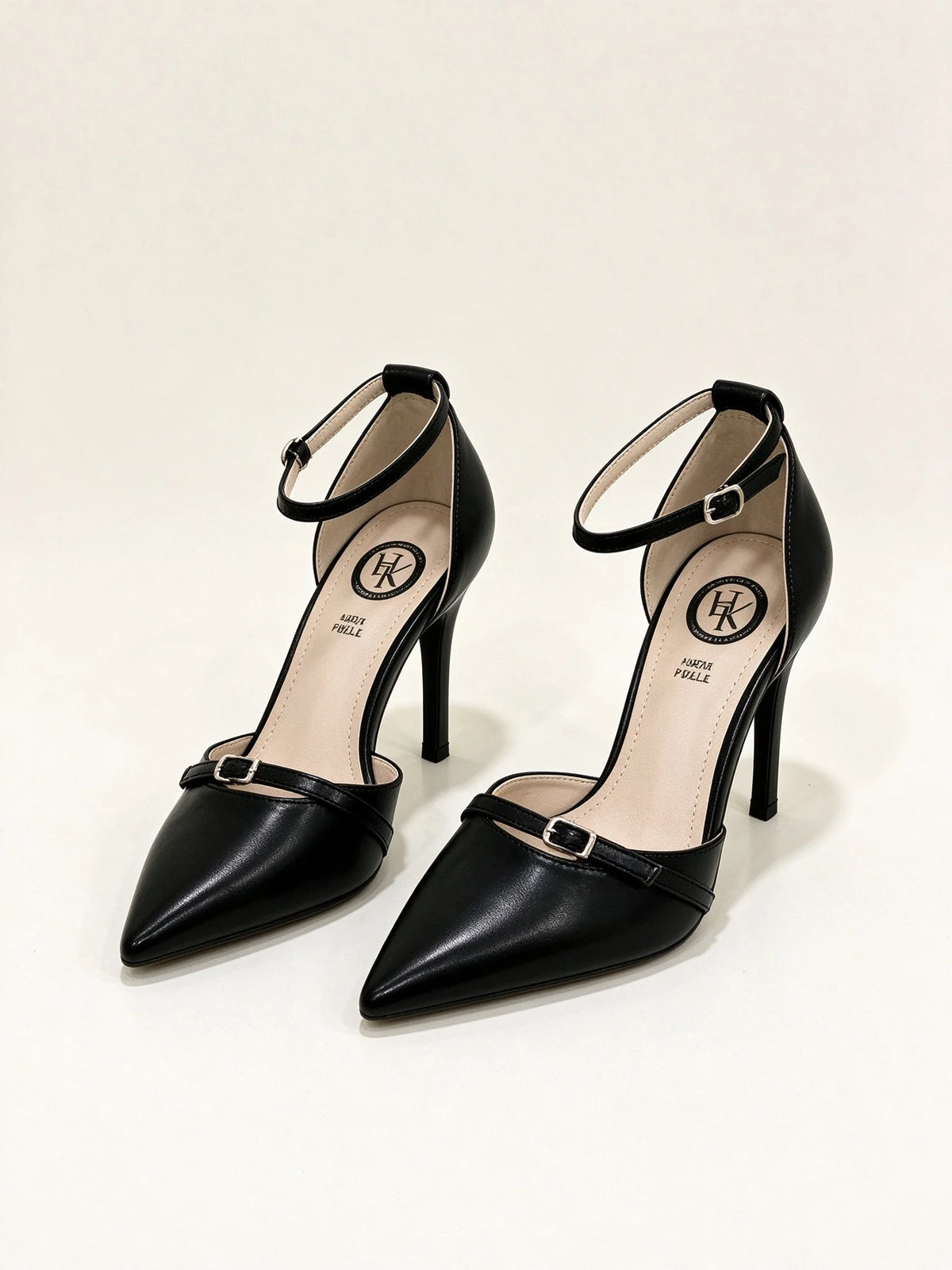 Un paio di scarpe décolleté nere a punta con tacco a spillo, cinturino alla caviglia e dettaglio fibbia sulla punta, su sfondo bianco - SUNTIMEMODA