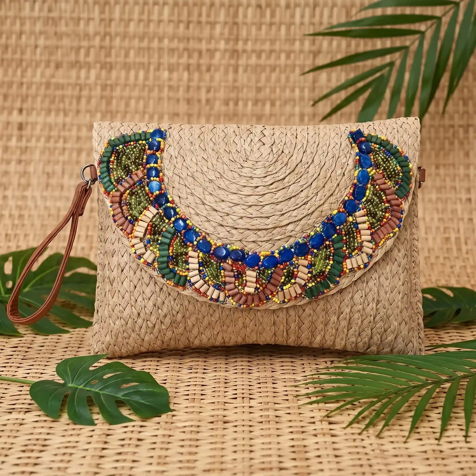 Pochette estiva in paglia intrecciata marrone chiaro con ricamo etnico di perline, posata su un tappeto di paglia con foglie tropicali - SUNTIMEMODA