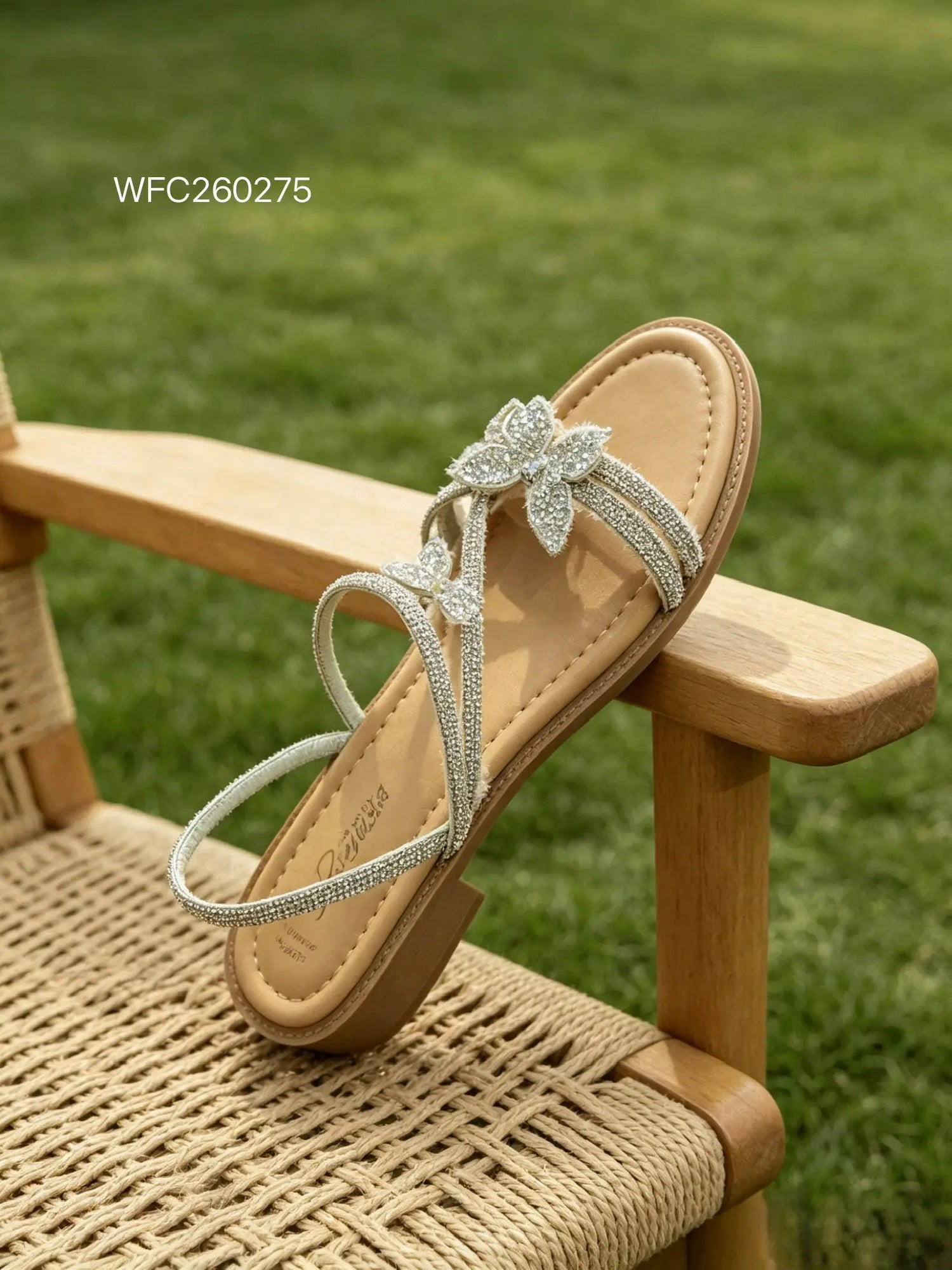 Sandalo piatto estivo da donna color beige e argento ricoperto di strass, con cinturino posteriore e due farfalle tridimensionali lucide, adagiato di lato su una sedia intrecciata in legno - SUNTIME MODA