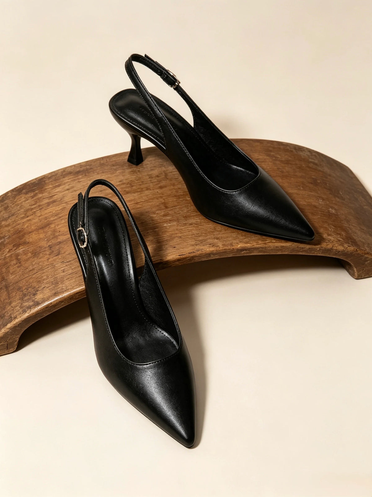 Un paio di décolleté slingback nere a punta con tacco a spillo, esposte elegantemente su un supporto in legno curvato - SUNTIMEMODA