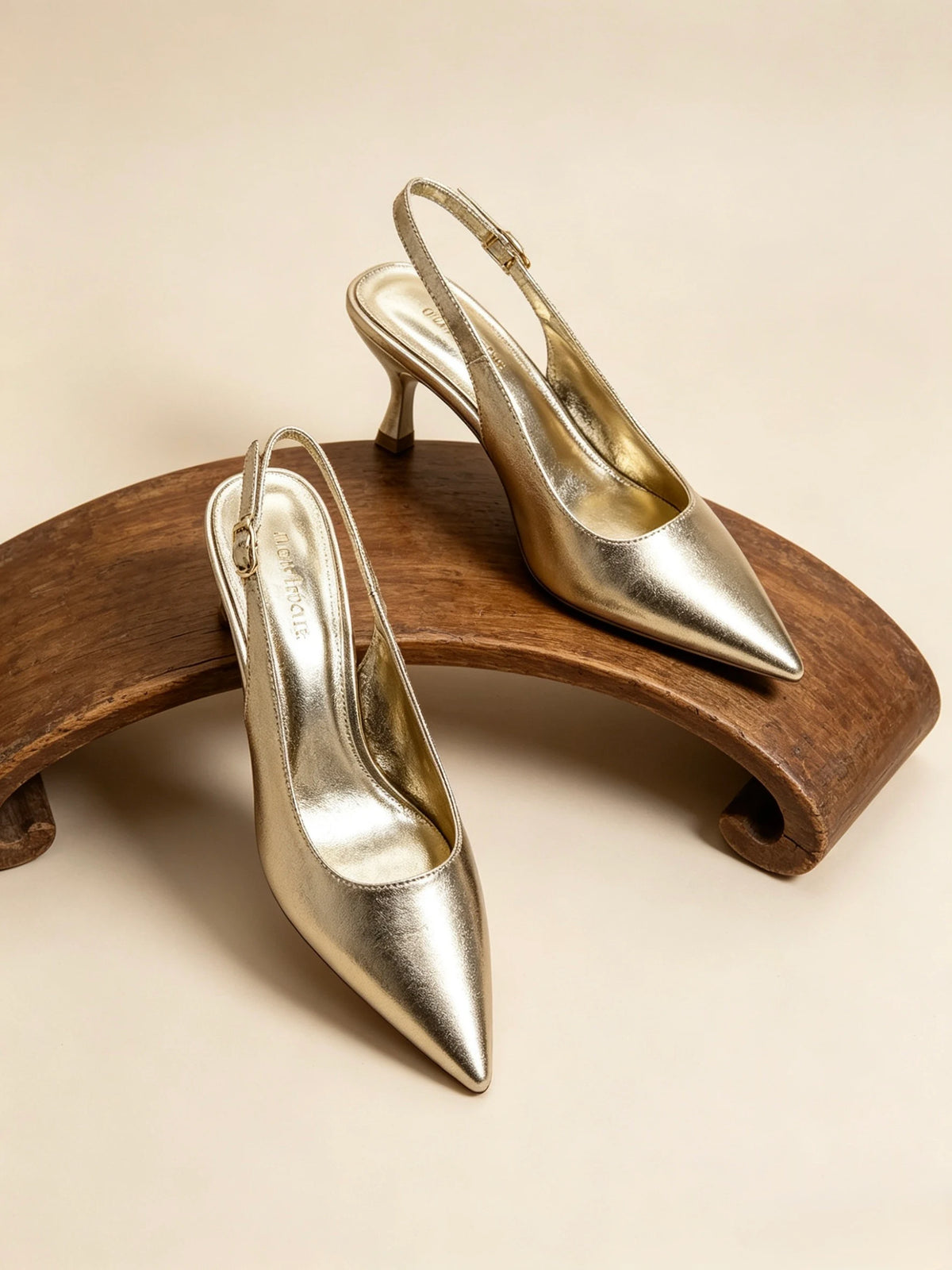 Un paio di luminose décolleté slingback color oro a punta con tacco a spillo, posizionate su un espositore in legno curvato - SUNTIMEMODA