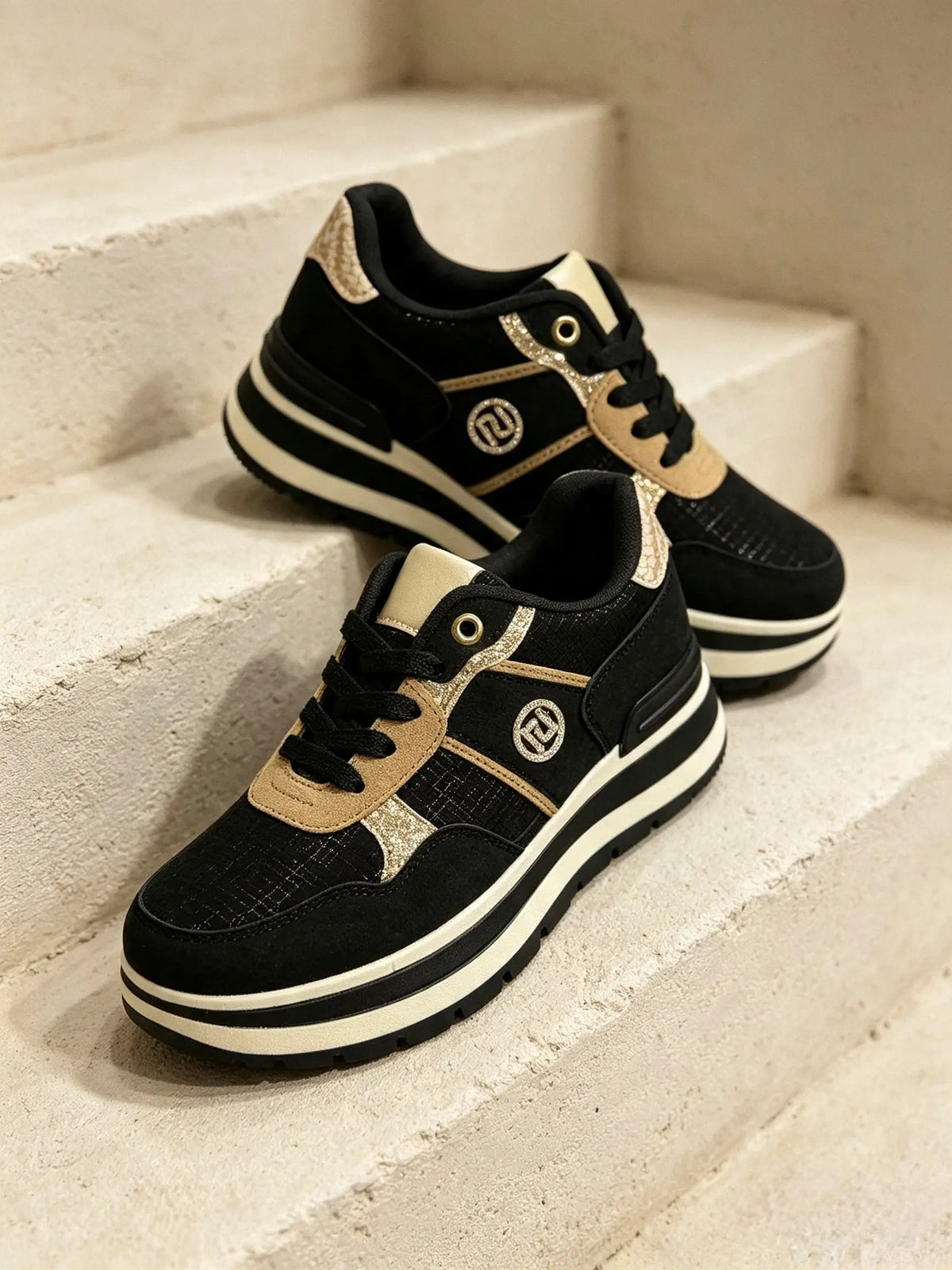 Un paio di sneakers chunky da donna JOMIX di colore nero, con inserti in tessuto e dettagli laminati color oro, posate su gradini in pietra - SUNTIMEMODA